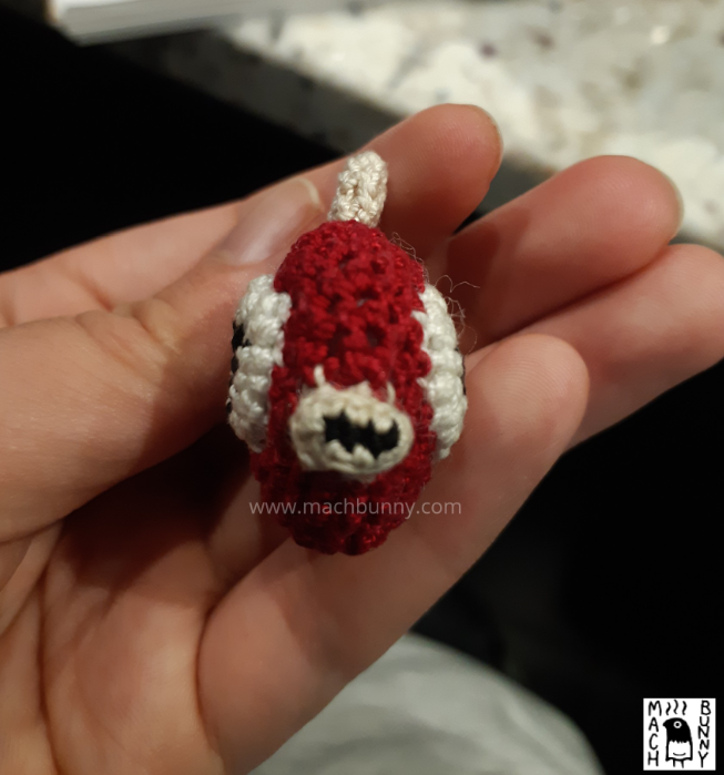 A small crocheted Magikarp, based off of the menu sprites from pokemon gold, silver, and crystal. The piece is facing forwards and resembles a small, round, red fish, with large eyes, small lips, and tan-coloured fins. It is smaller than the person's three fingers, where it rests on top.